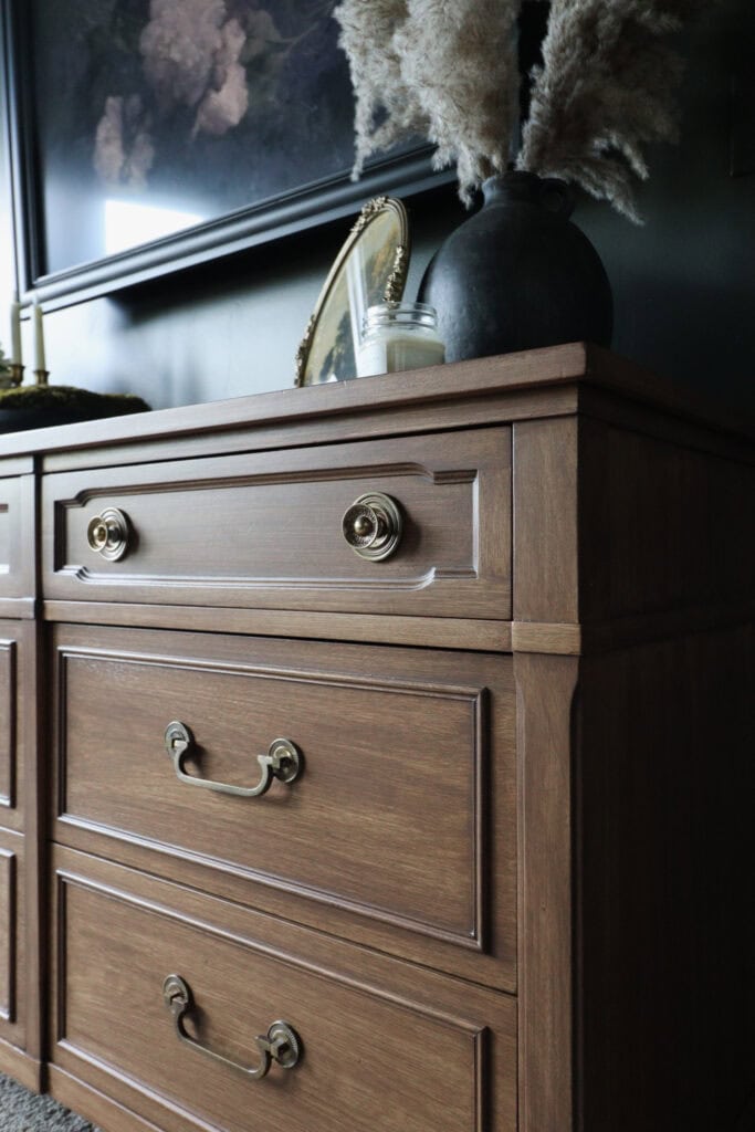 Wooden dresser with gold handles, and candle, frame, and vase on top.
