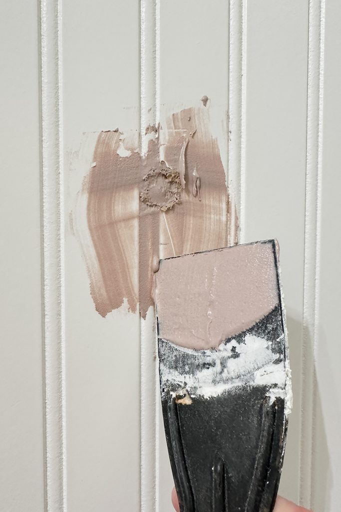 A putty knife spreads filler over a hole in a white, grooved wall for repair.