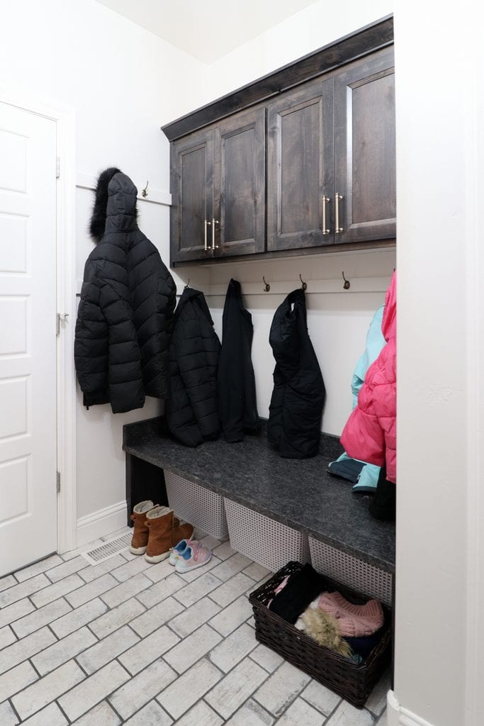 Coats hang on hooks above a bench; shoes and a basket with gloves are below in a tiled entryway.
