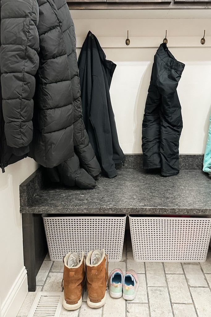 Three dark coats hang above a gray bench with baskets, boots, and colorful shoes on a tiled floor.