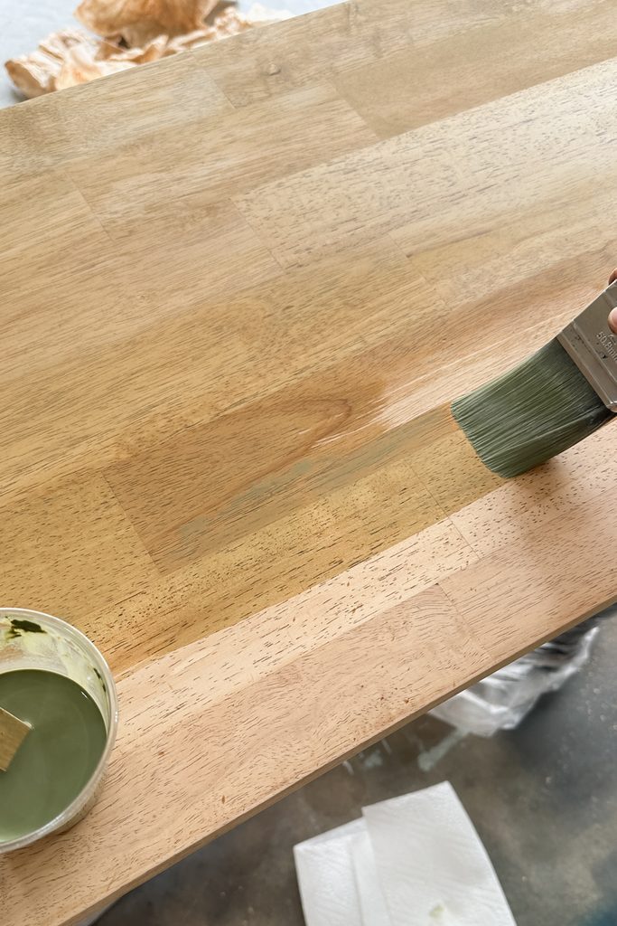 A hand uses a brush to apply green paint wash to a wooden surface