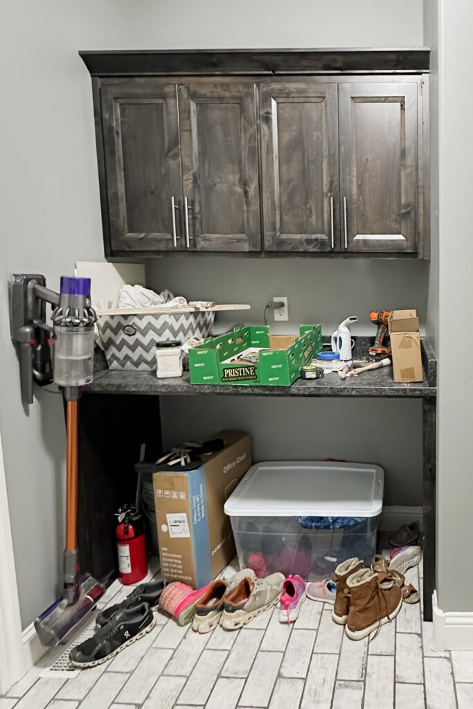 Mudroom shelf with cleaning supplies, boxes, shoes, and storage bins below a dark wood cabinet.