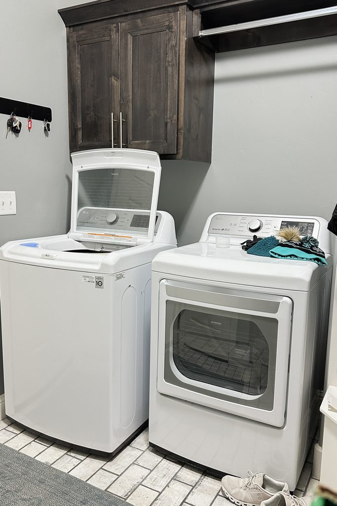 Washing machine and dryer in laundry room, cabinet above, clothes on dryer, shoes on the floor.