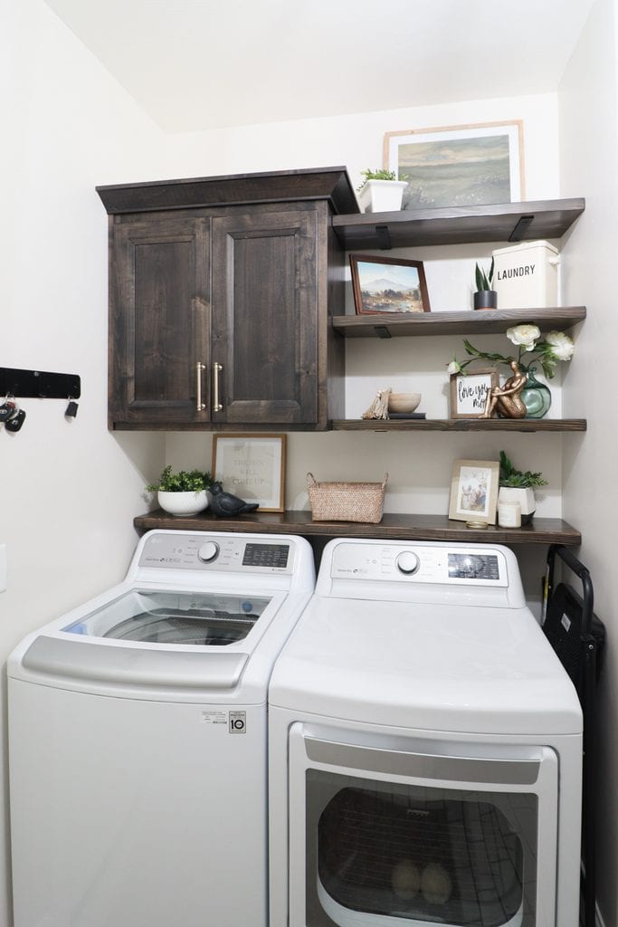 A laundry room with a washer, dryer, dark cabinets, shelves, plants, baskets, and decor items.