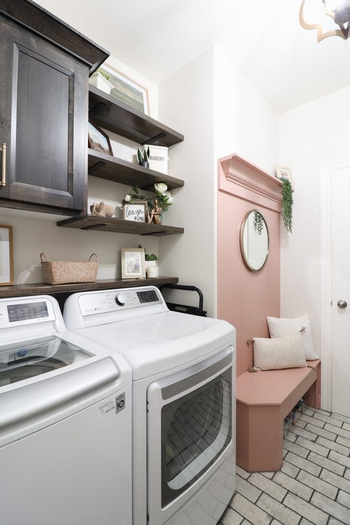 Laundry room with white washer and dryer, wooden shelves, pink wall and bench, and decorative items on display.