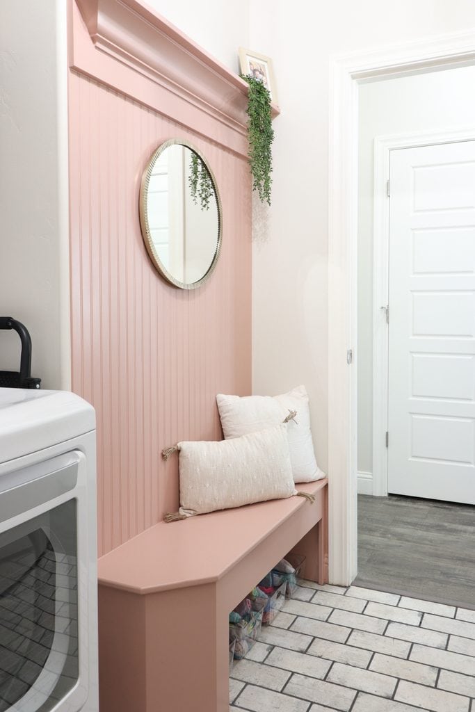 Pink bench with pillows, round mirror, hanging plant, and a white dryer in a bright laundry room.