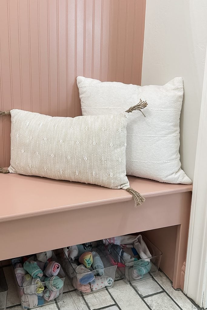 Two white pillows on a pink bench with clear bins of rolled socks underneath on a tiled floor.