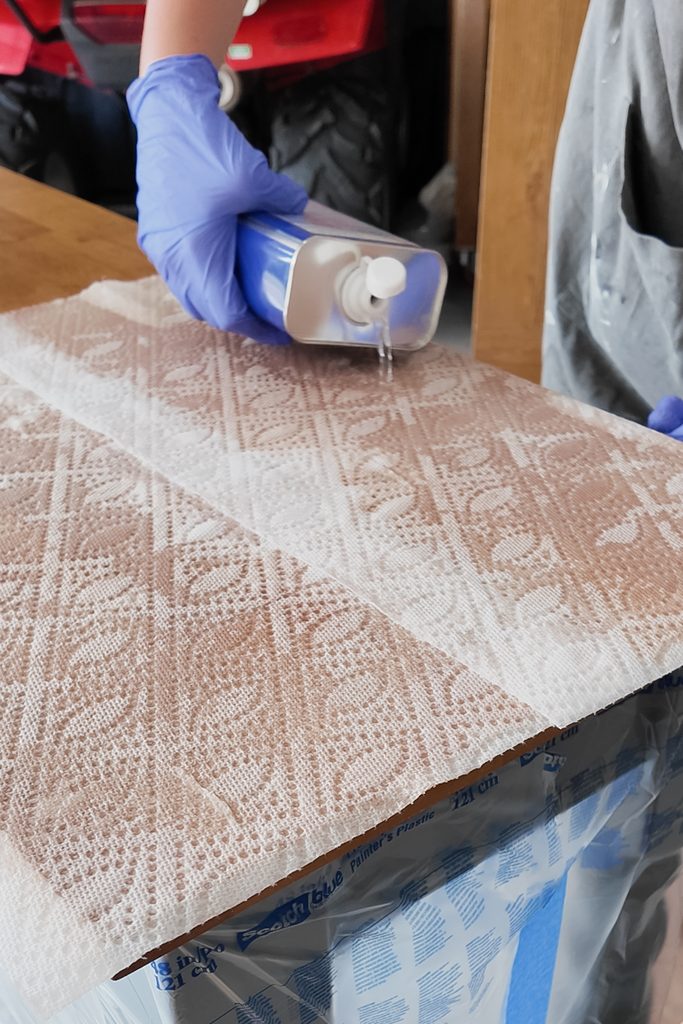 A gloved hand pours acetone from a can onto paper towels on table top