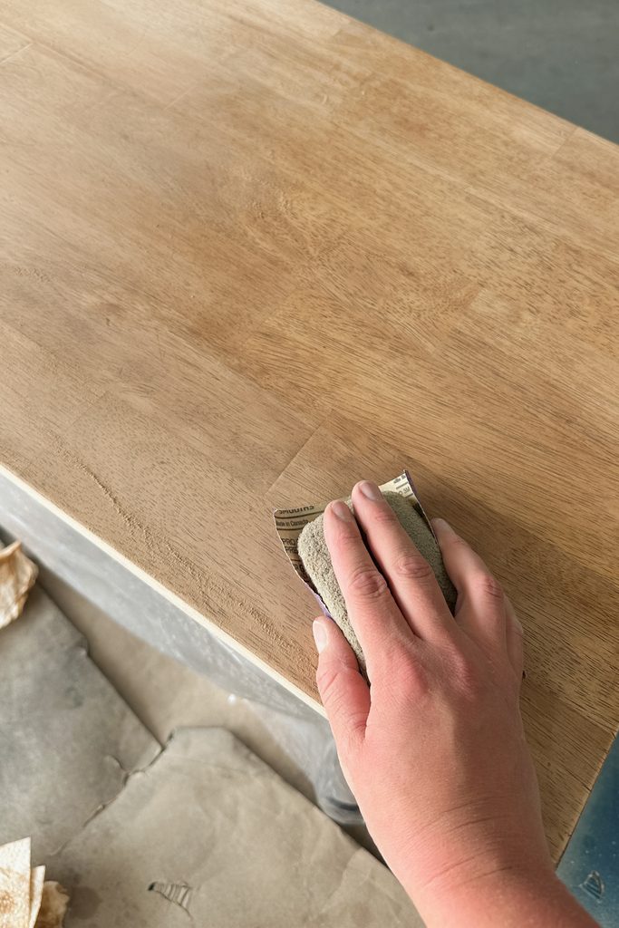 A hand sands a wooden surface with sandpaper and sanding block, smoothing the wood.
