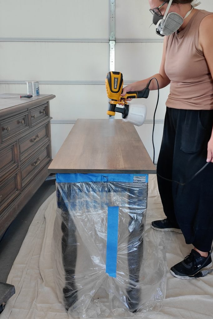 Person wearing mask uses a paint sprayer to finish a wooden table in a garage