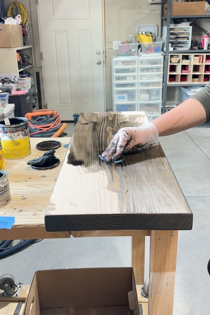 A gloved hand applies dark wood stain to a wooden plank in a workshop with tools and supplies.