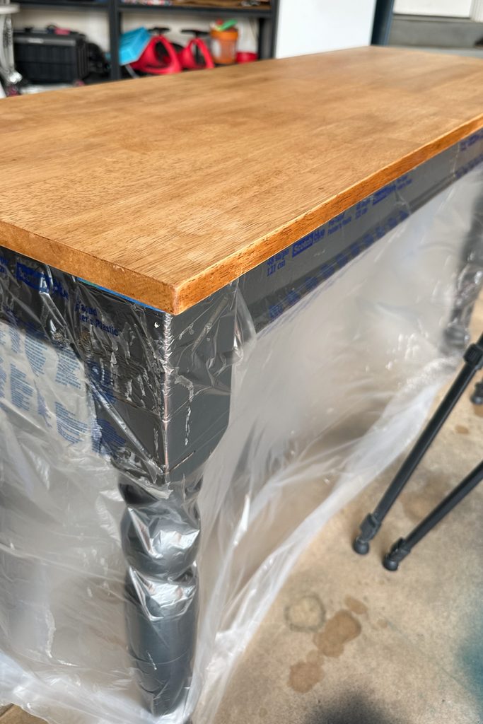 A wooden table with a finished top, legs wrapped in plastic for protection, stands in a garage.