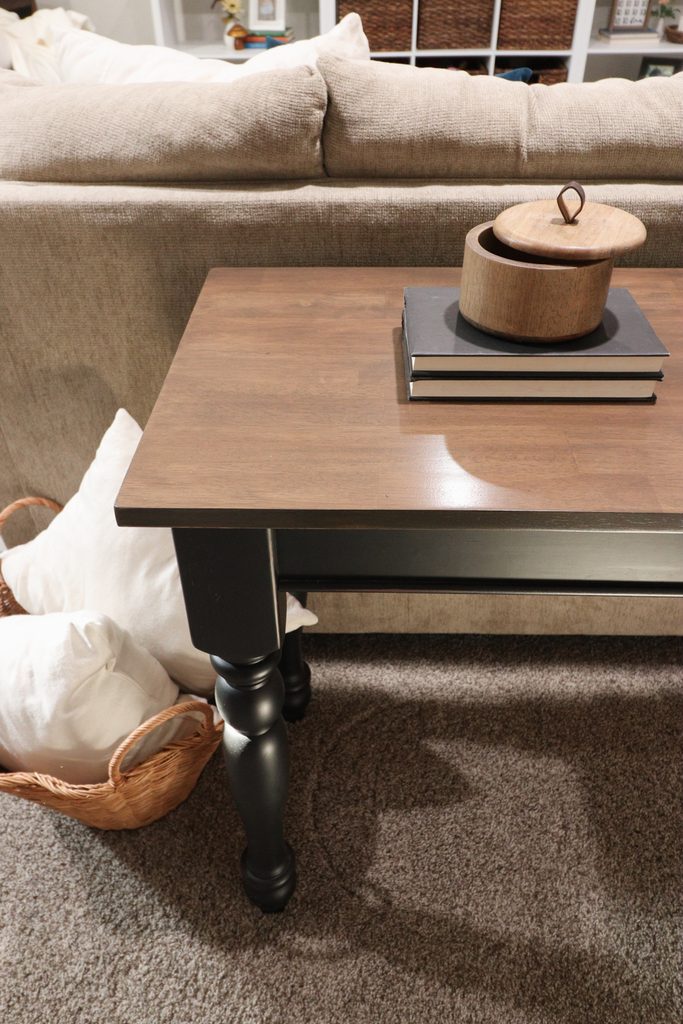 Wooden table with stacked books and a round wooden basket beside a beige sofa on a carpeted floor.