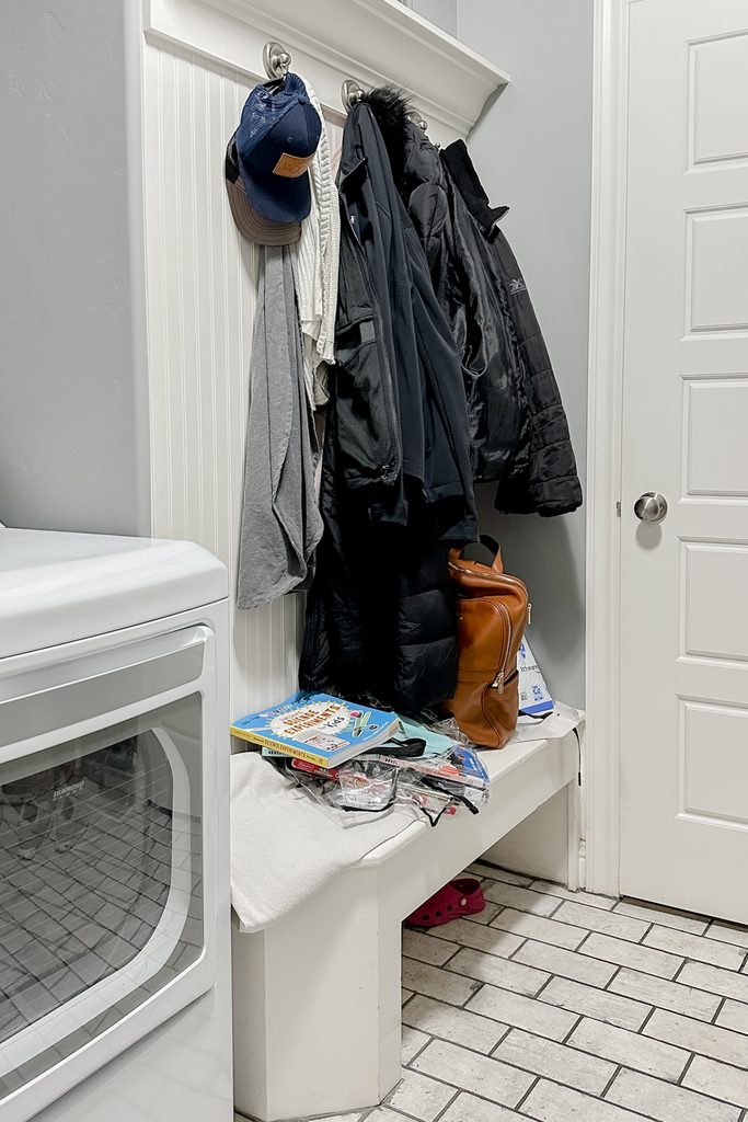 Coats and hats hang on hooks above a bench with bags, papers, and laundry items in a mudroom.