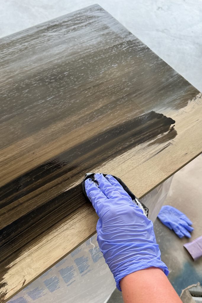 A gloved hand applies dark stain to a wooden surface with a cloth