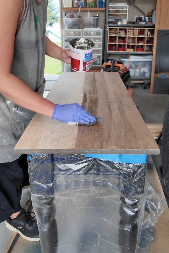 Person staining a wooden tabletop in a workshop while wearing blue gloves and holding a can of wood stain.