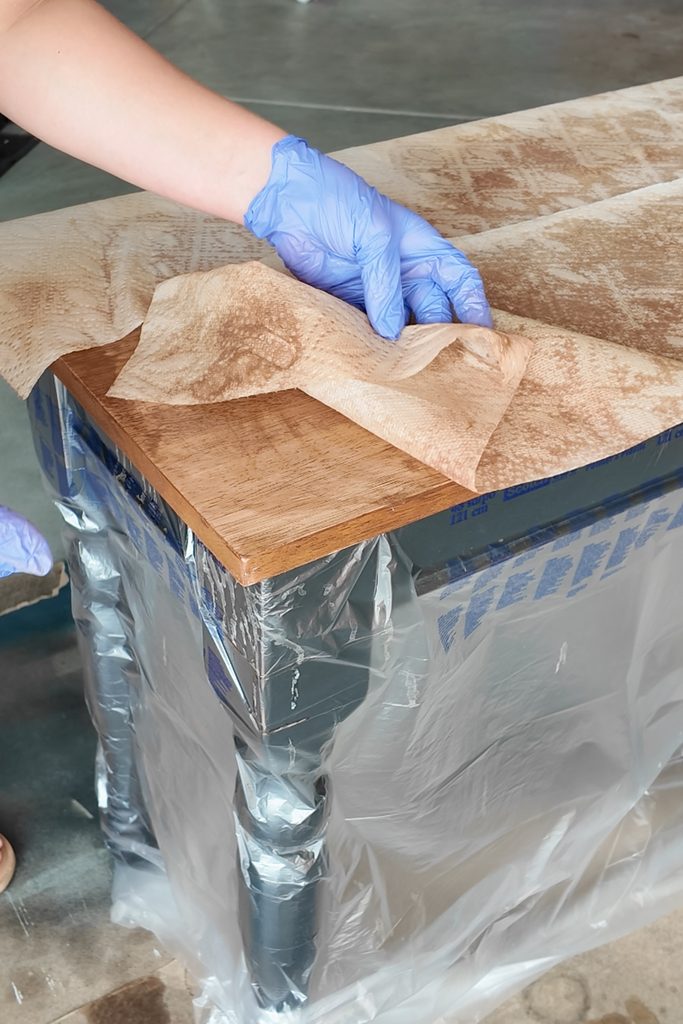 Gloved hand wiping old finish off a wooden table top, which is partially covered in plastic sheeting.