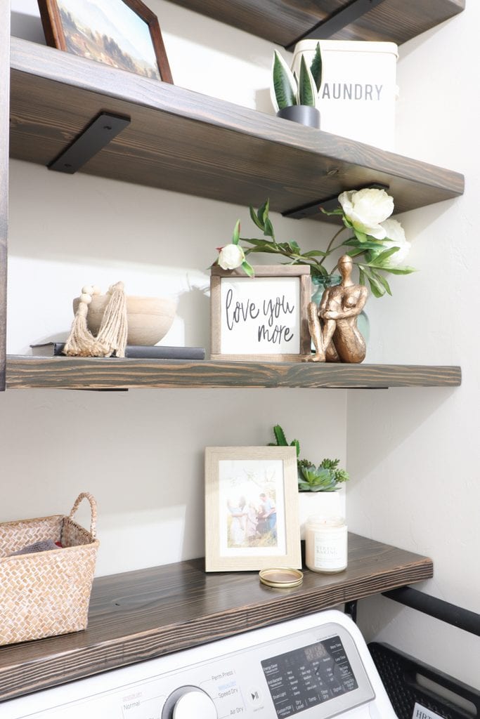 Wood shelves over a washer hold decor: plants, framed photos, a Love You More sign, and baskets.
