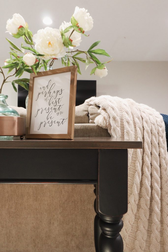 Table with a framed quote, white flowers in a vase, and a cream knit blanket draped over the back.