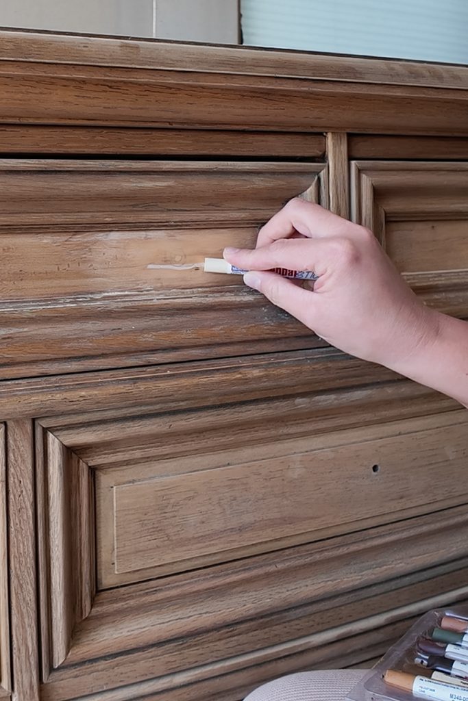 A hand uses a wax filler stick to repair a scratch on a wooden dresser drawer.