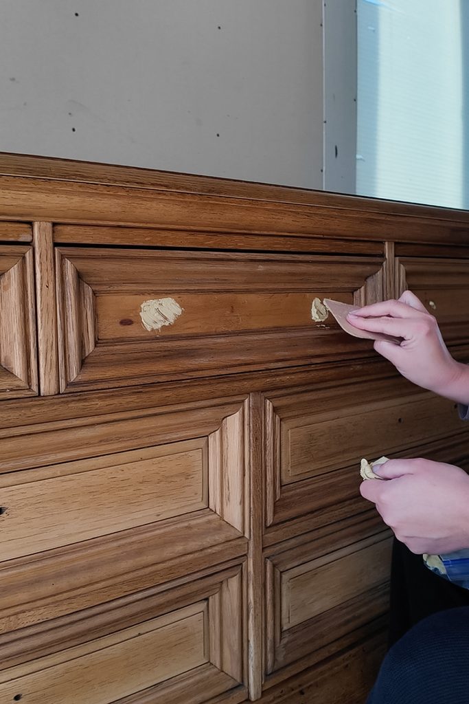 A person uses a putty knife to apply wood filler to a wooden dresser drawer.