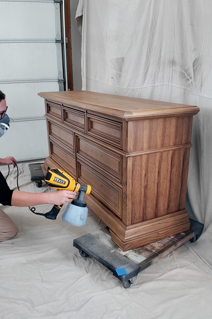 A person wearing a respirator uses a paint sprayer on a wooden dresser.