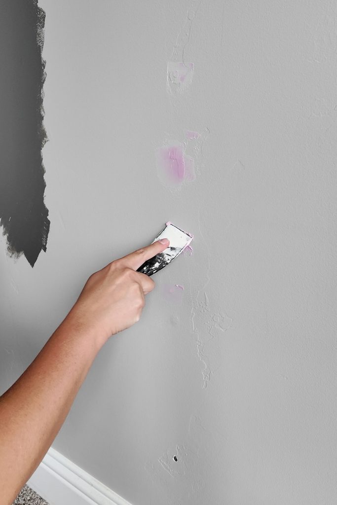 Hand using a putty knife to apply pink spackle to a gray wall.