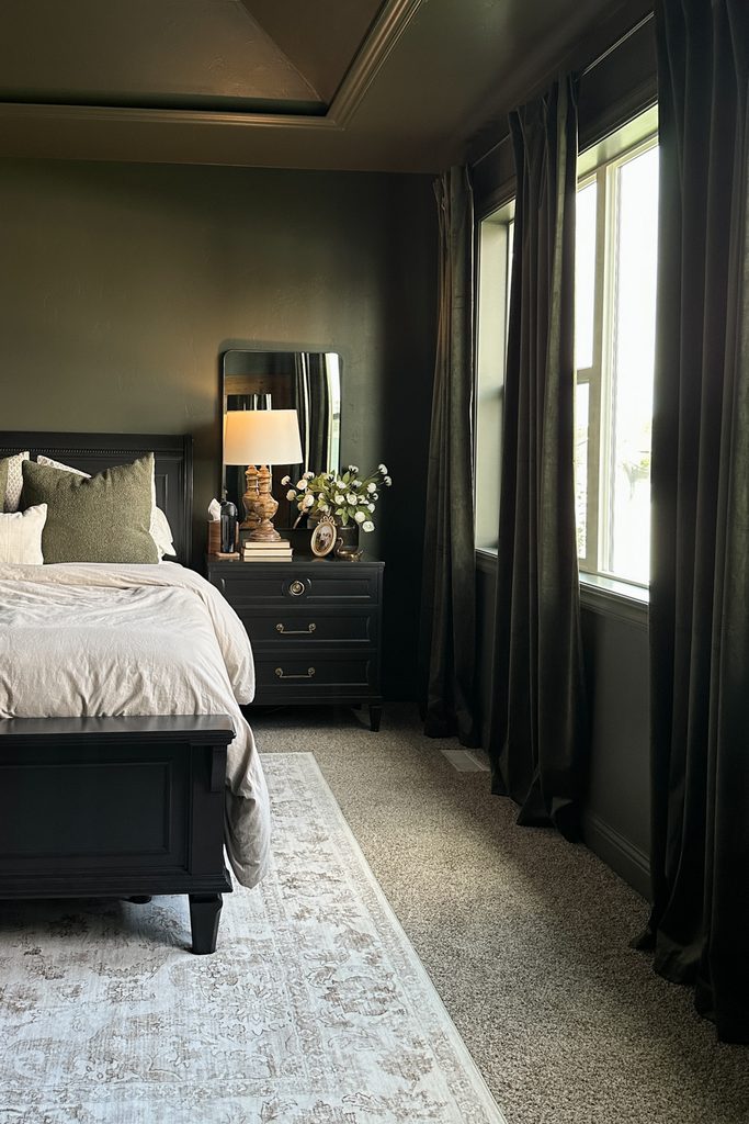 A dark green color drenched bedroom with a black bed, a nightstand holding a lamp and flowers, and floor-length velvet curtains.