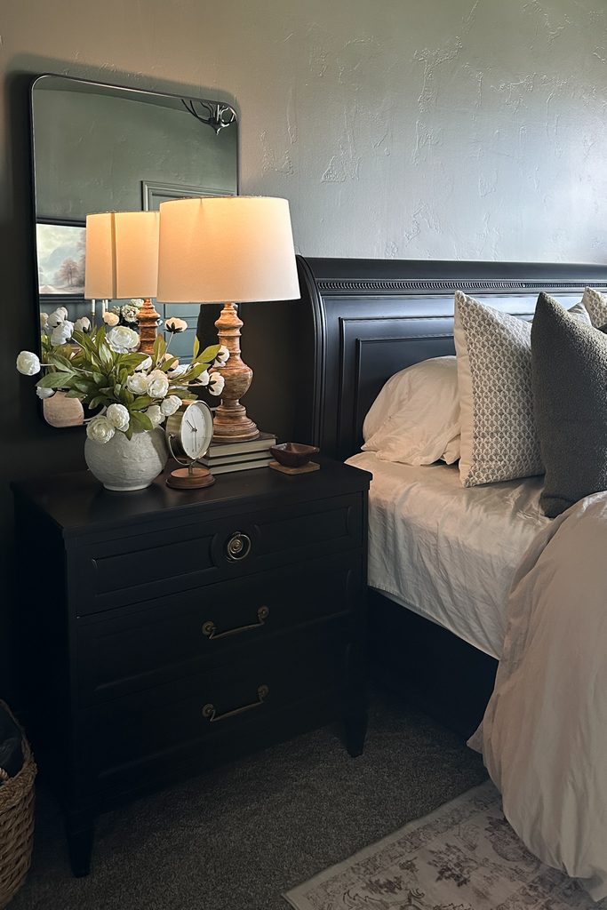 A black nightstand next to a bed with a lamp, a vase of white flowers, and a small clock in a dark green color drenched bedroom