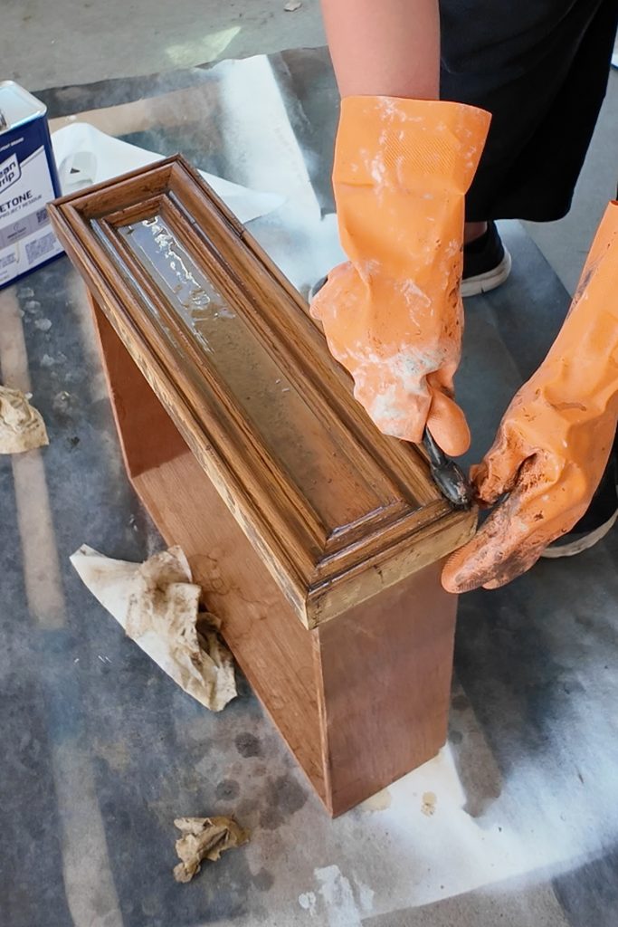 Person wearing orange rubber gloves uses a brush to strip finish from a wooden drawer front.