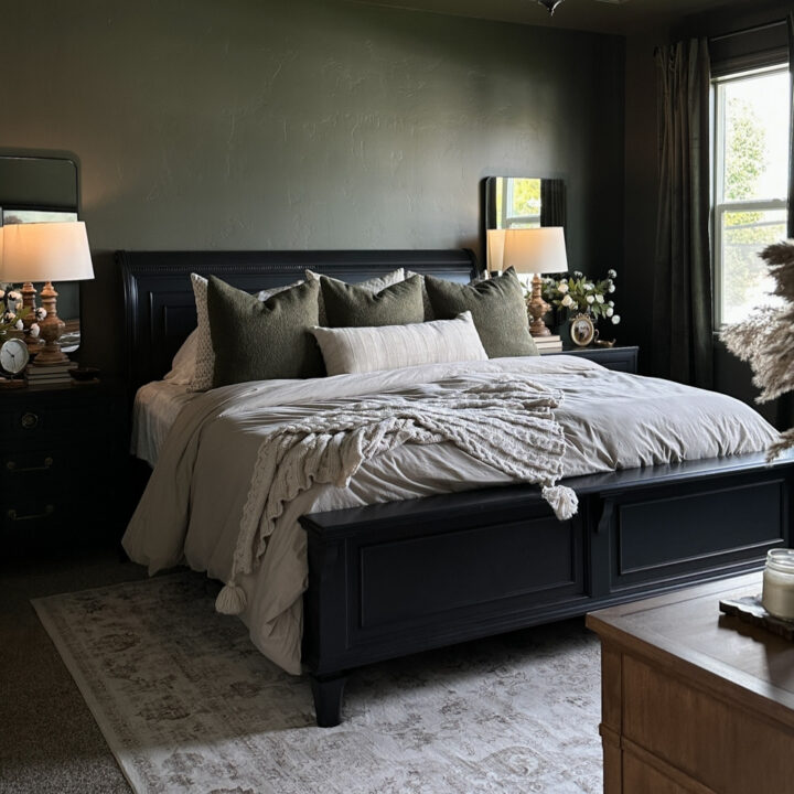 A black wooden bed with olive green and cream pillows in a bedroom with dark green walls.