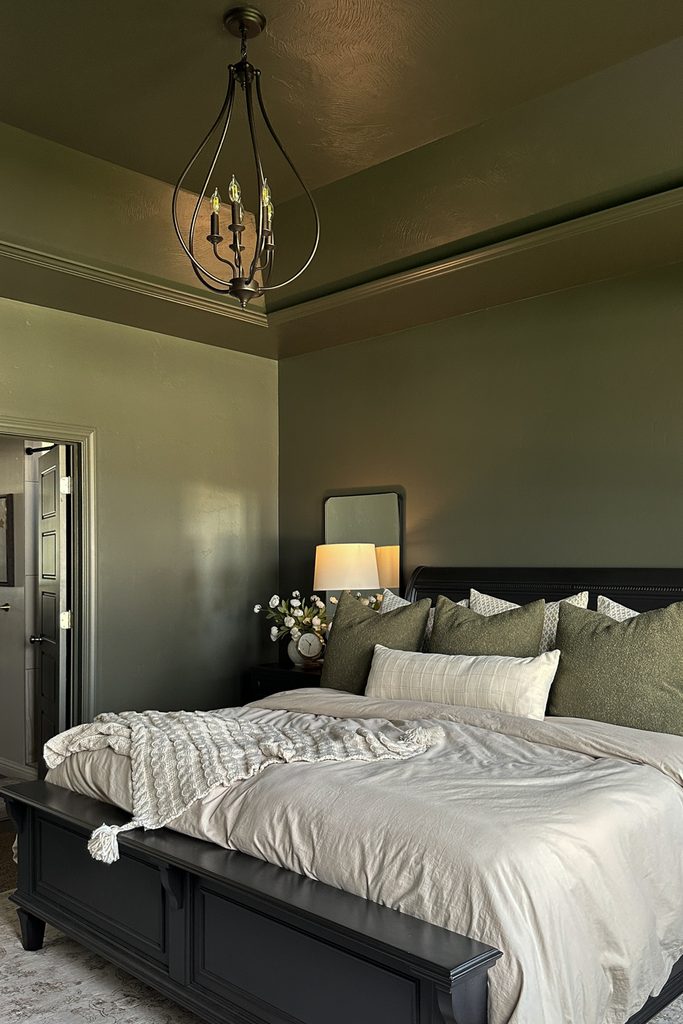 A dark green painted bedroom with a black bed frame, cream bedding, and a hanging lantern style chandelier.