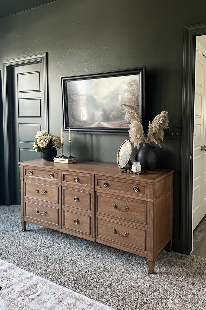 A wooden dresser against a dark green wall, topped with a DIY framed TV with landscape painting and decorative items.