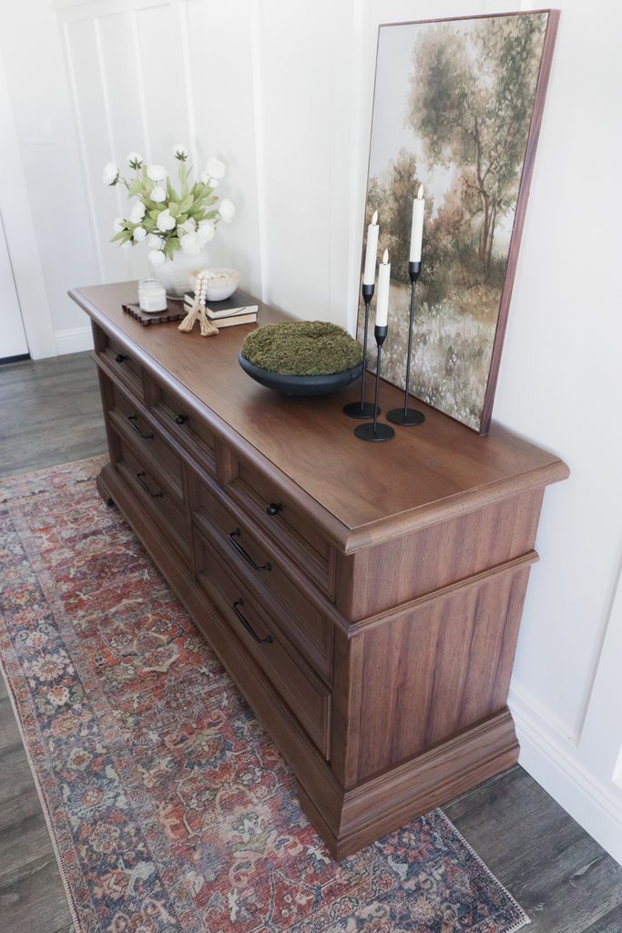 Wooden dresser topped with a vase of flowers, a bowl of moss, candles, and framed landscape art.