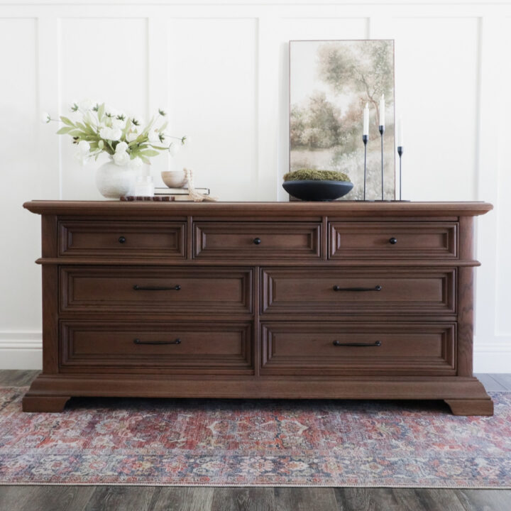 Dark wood dresser with seven drawers and black hardware, topped with a vase of flowers, a bowl, and framed art.