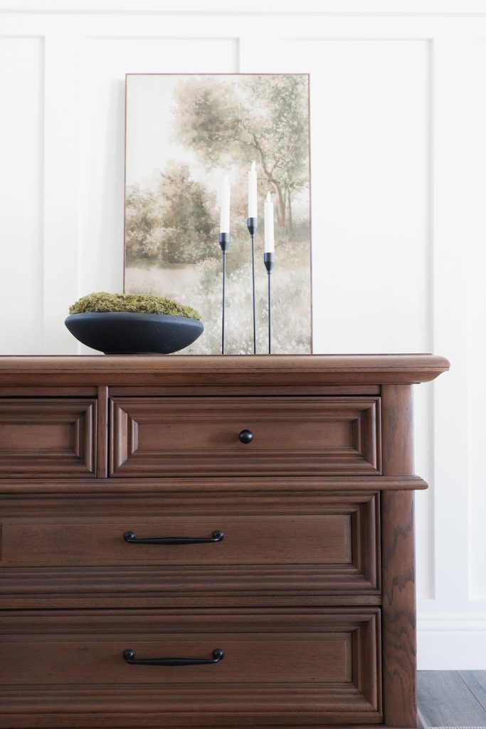 A wooden dresser topped with a bowl of moss and three candle holders in front of a landscape painting.