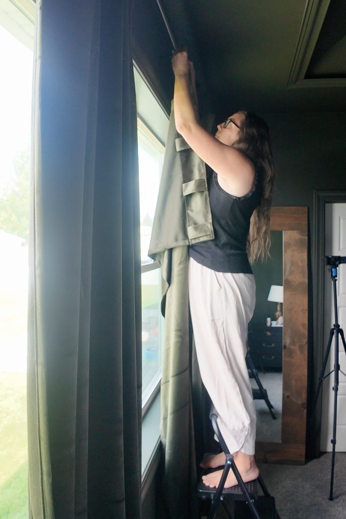 A person stands on a step stool to hang a curtain on a rod.