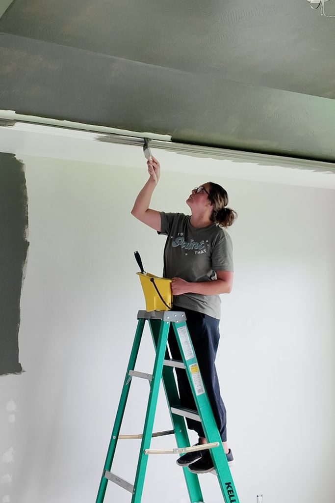 A person on a stepladder uses a paintbrush to paint the edge of a ceiling.