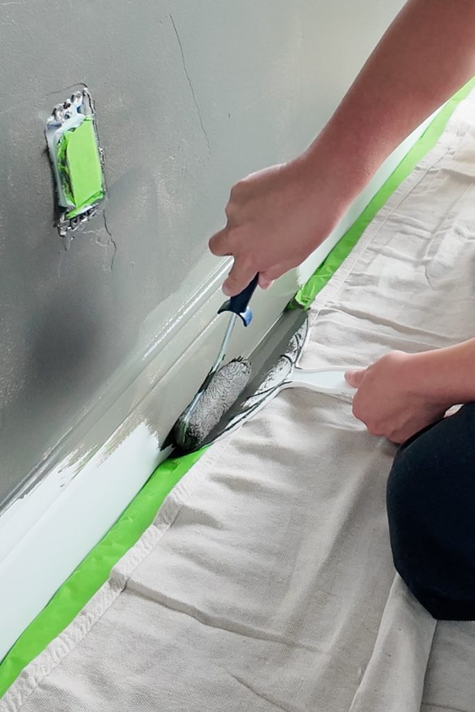 A person uses a small paint roller to paint a baseboard without getting paint on the carpet.