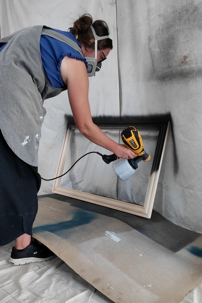 A person wearing a respirator uses a paint sprayer to coat a wooden picture frame with black paint.