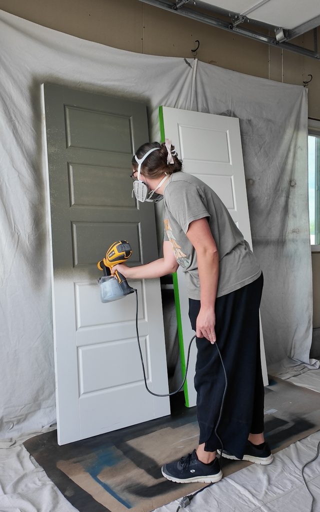 A person wearing a respirator uses a paint sprayer to paint a white door dark green.