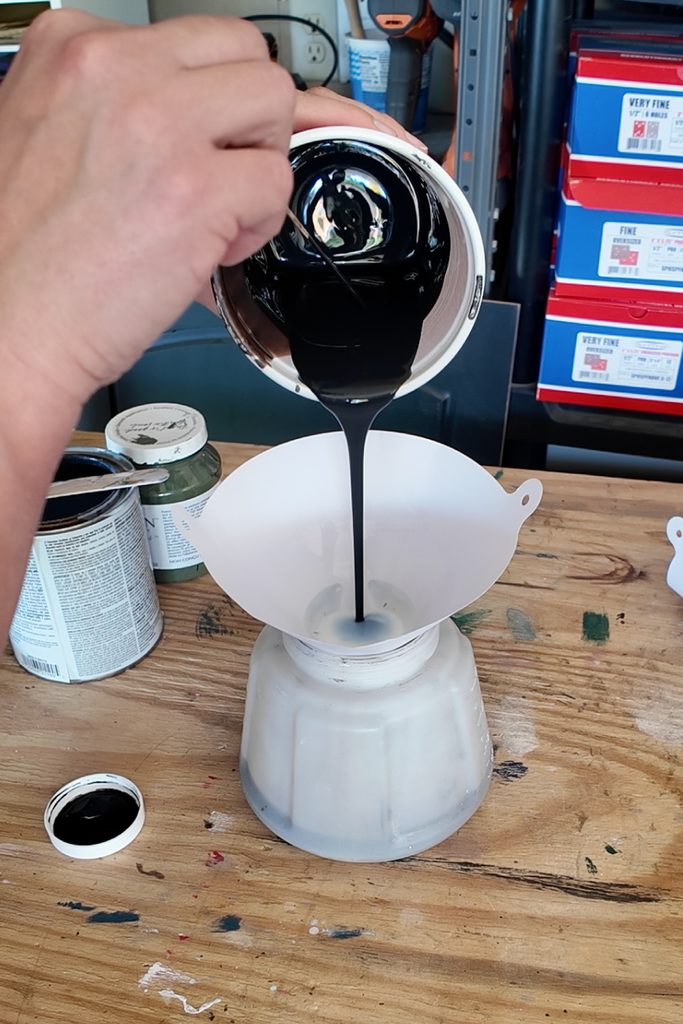 A hand pours black paint from a container into a white filter resting on a sprayer container