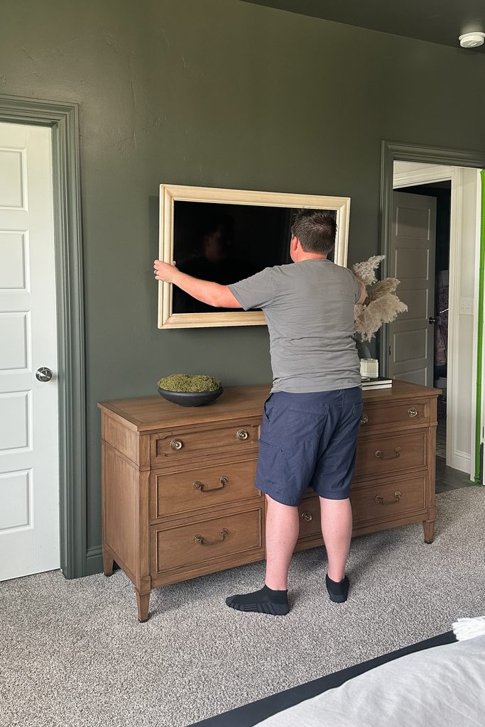 A person hanging a DIY frame onto a TV with a dark green wall above a wooden dresser.