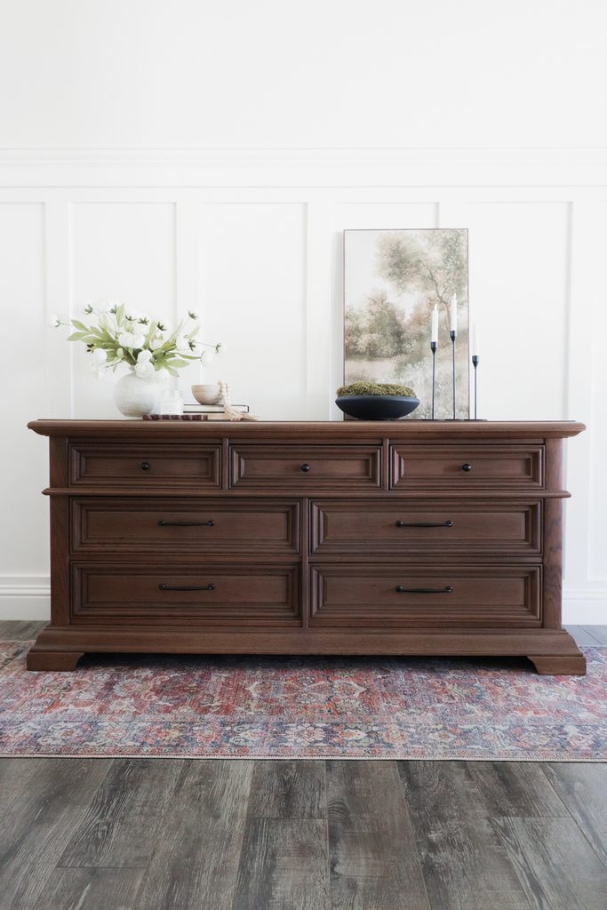 A dark wood dresser with seven drawers, topped with a vase of flowers, a framed landscape print, and candles.