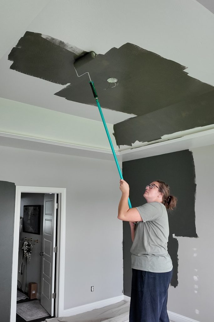 A person using a paint roller on a broom stick to paint a ceiling dark green.