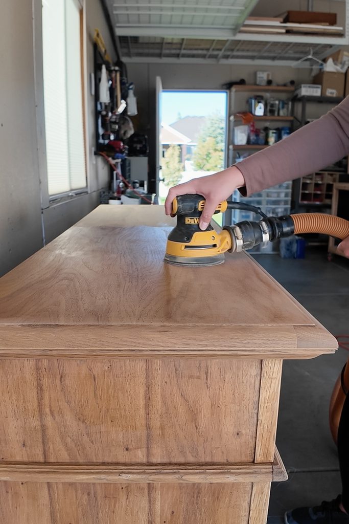 A person using a handheld power sander on the top of a wooden dresser.
