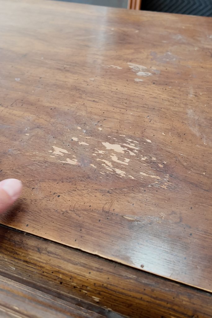 A finger points to a patch of worn finish and scratches on a wooden table surface.