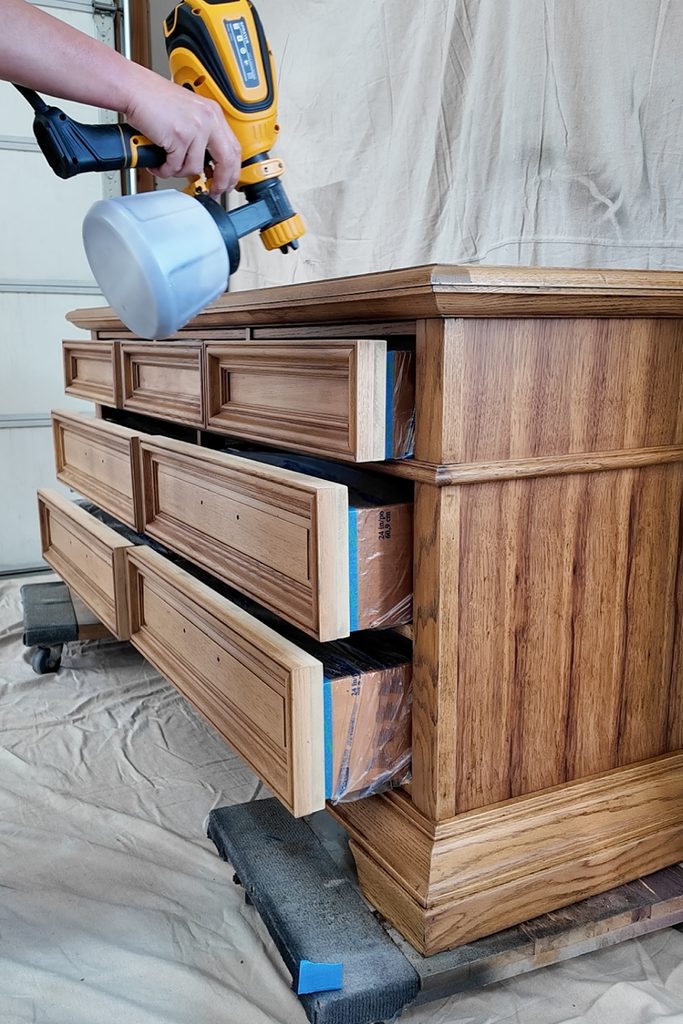 A person uses a yellow paint sprayer on a wooden dresser with open drawers.