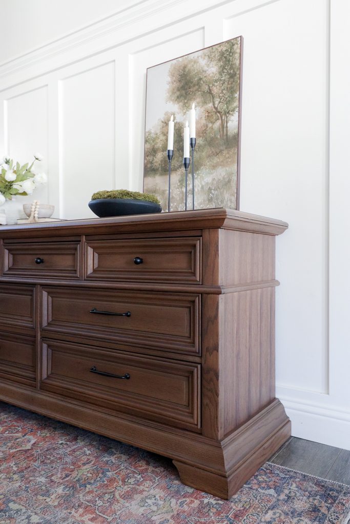 Dark wood dresser with a landscape painting, candles, and a moss bowl on top.