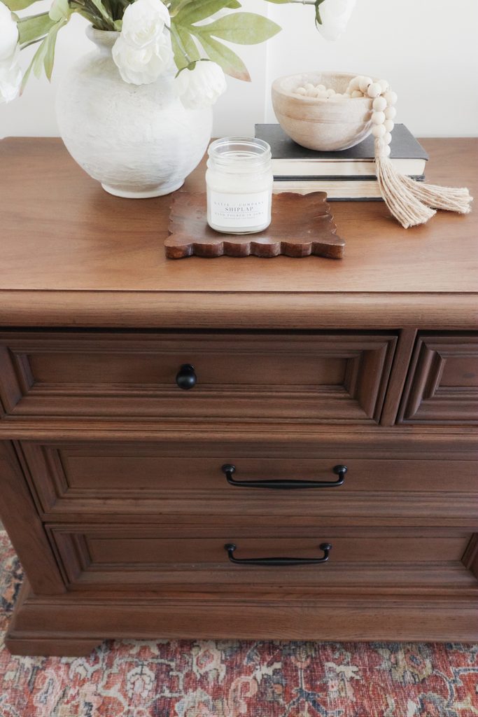 A wooden dresser topped with a vase of flowers, a scented candle, books, and a decorative bowl with wood beads.